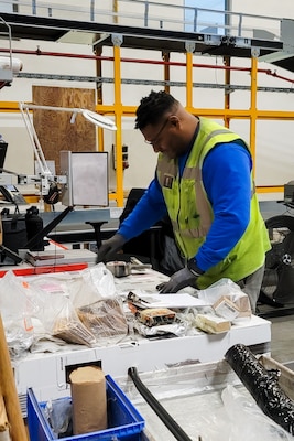 A man in a reflective safety vest looks through shipping labels and receipts at a table in a warehouse.