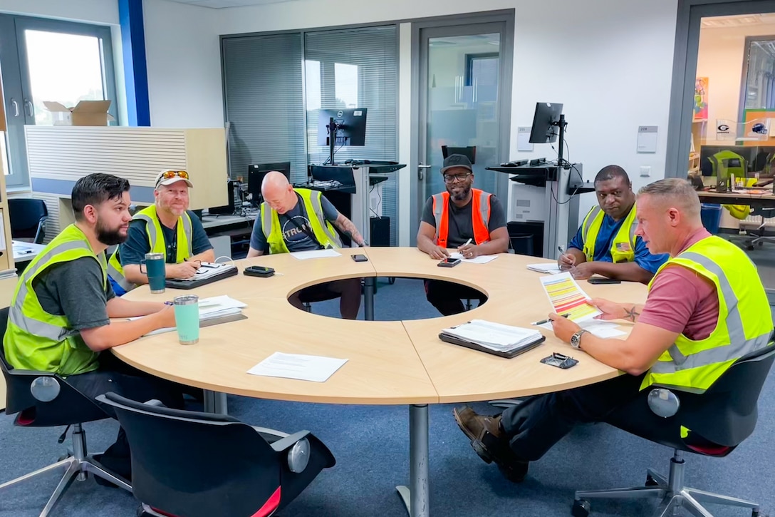 A group of people wearing reflective safety vests sit around a round conference table talking.