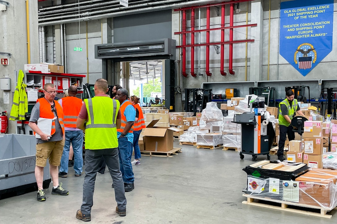 A group of people wearing reflective safety vests stand talking in a warehouse with boxes and pallets in the background.