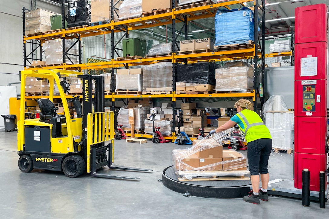 A woman in a reflective safety vest wraps a pallet of boxes with plastic wrap in a warehouse.