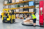 A woman in a reflective safety vest wraps a pallet of boxes with plastic wrap in a warehouse.