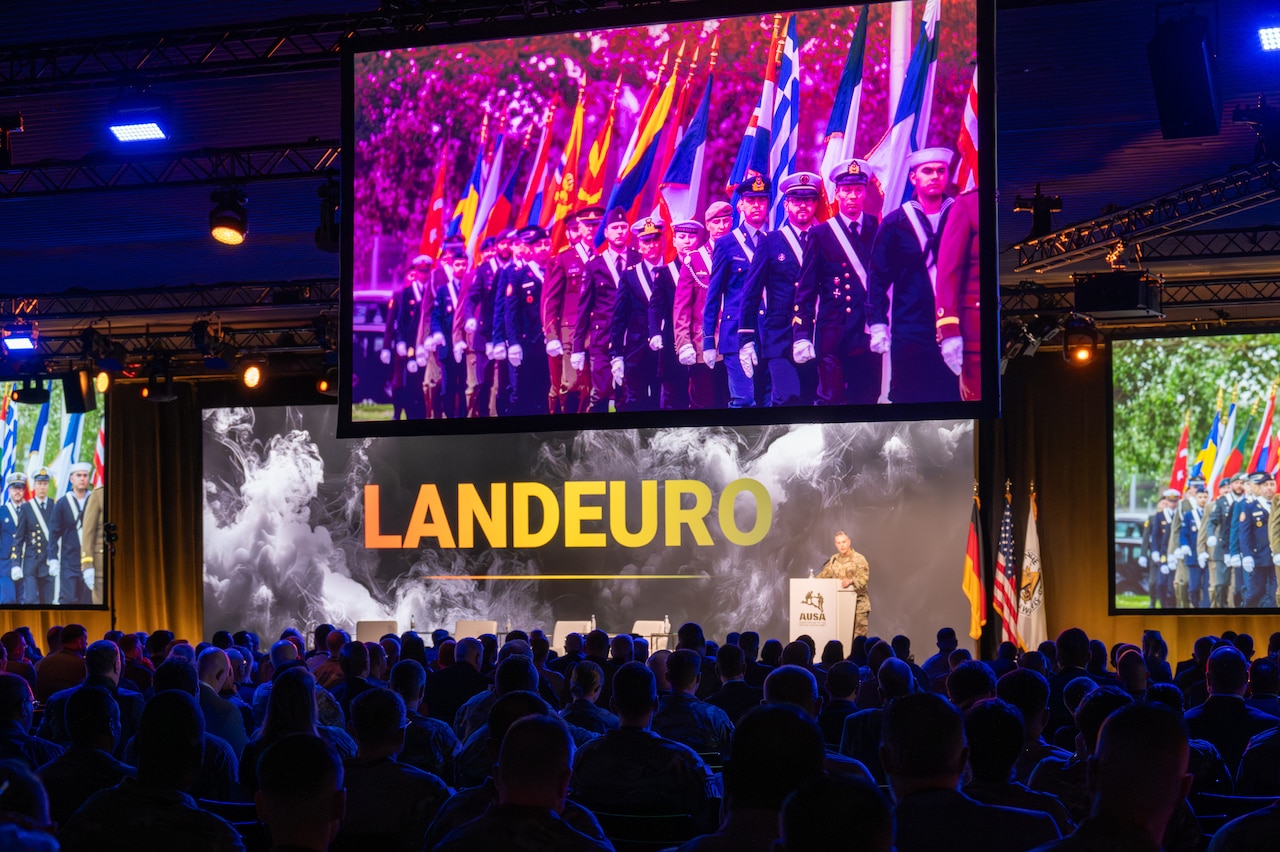 A large video screen hangs from a ceiling and shows photos of service members carrying flags. Below the screen the word "LANDEURO" is emblazoned on the wall. A man stands behind a lectern, in front of a large audience.