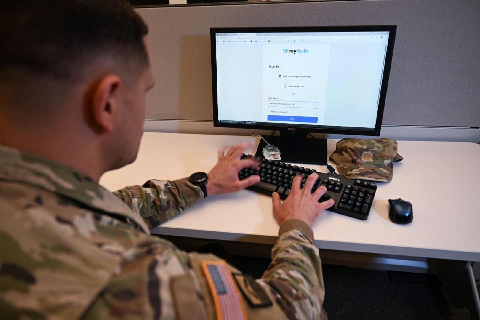 Uniformed service member types on keyboard in front of desktop showing myAuth login screen.
