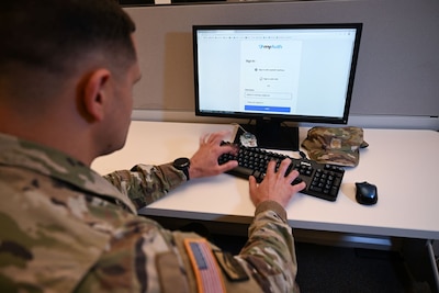 Uniformed service member types on keyboard in front of desktop showing myAuth login screen.
