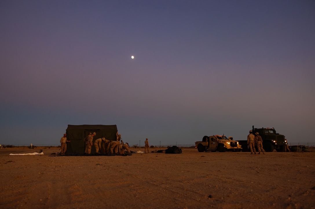 Marines set up a tent during lowlight while on a dessert terrain field.