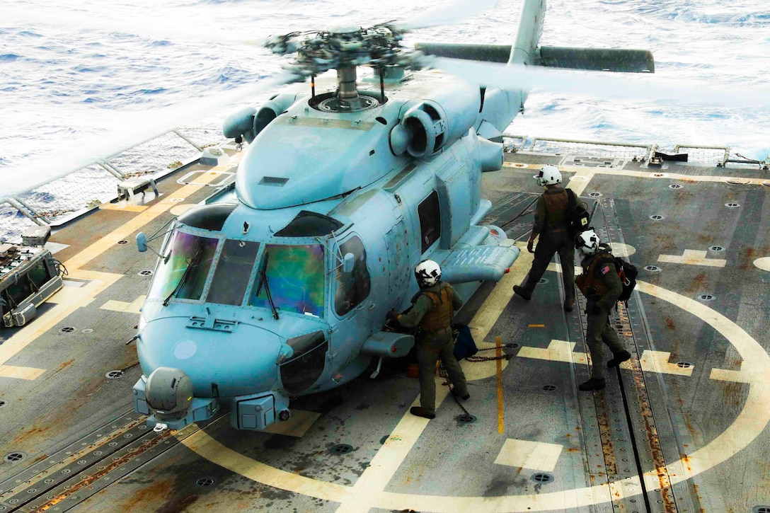 Three sailors with flight equipment move next to a helicopter parked on the deck of a military ship at sea.