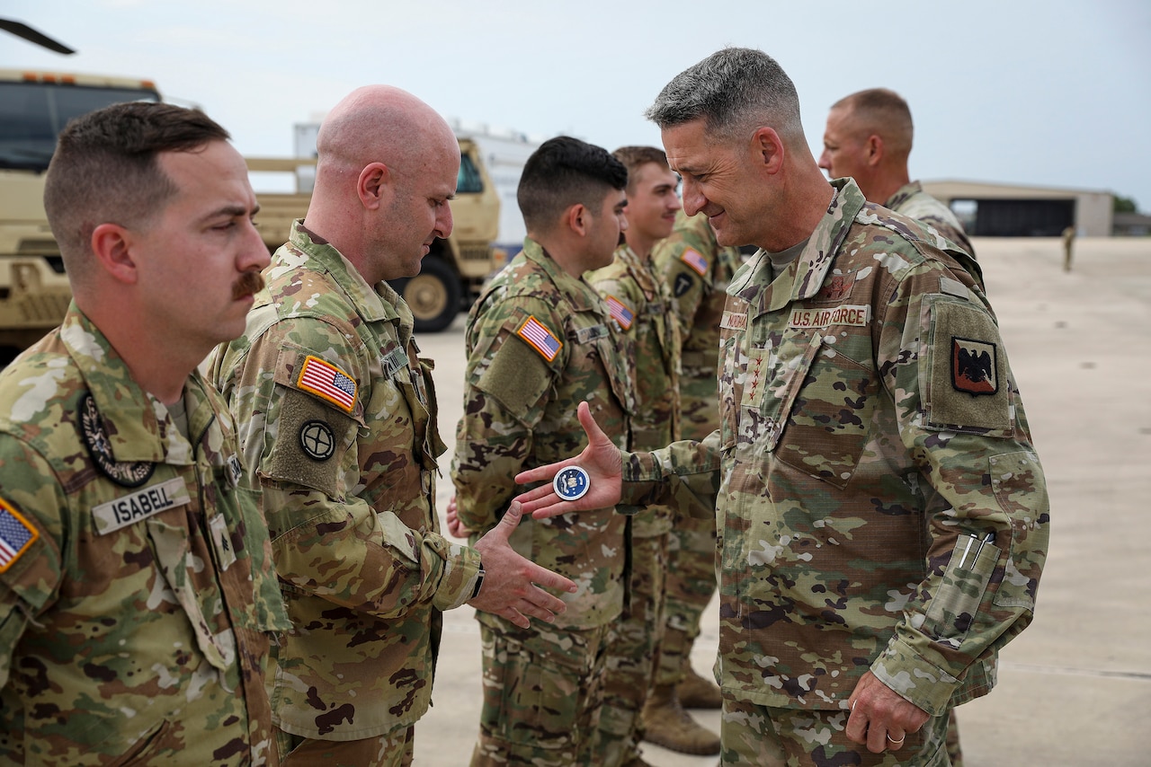 A man dressed in a military uniform presents a challenge coin to another man also dressed in a military uniform as four other men in camouflage uniforms stand in a line.