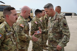 A man dressed in a military uniform presents a challenge coin to another man also dressed in a military uniform as four other men in camouflage uniforms stand in a line.