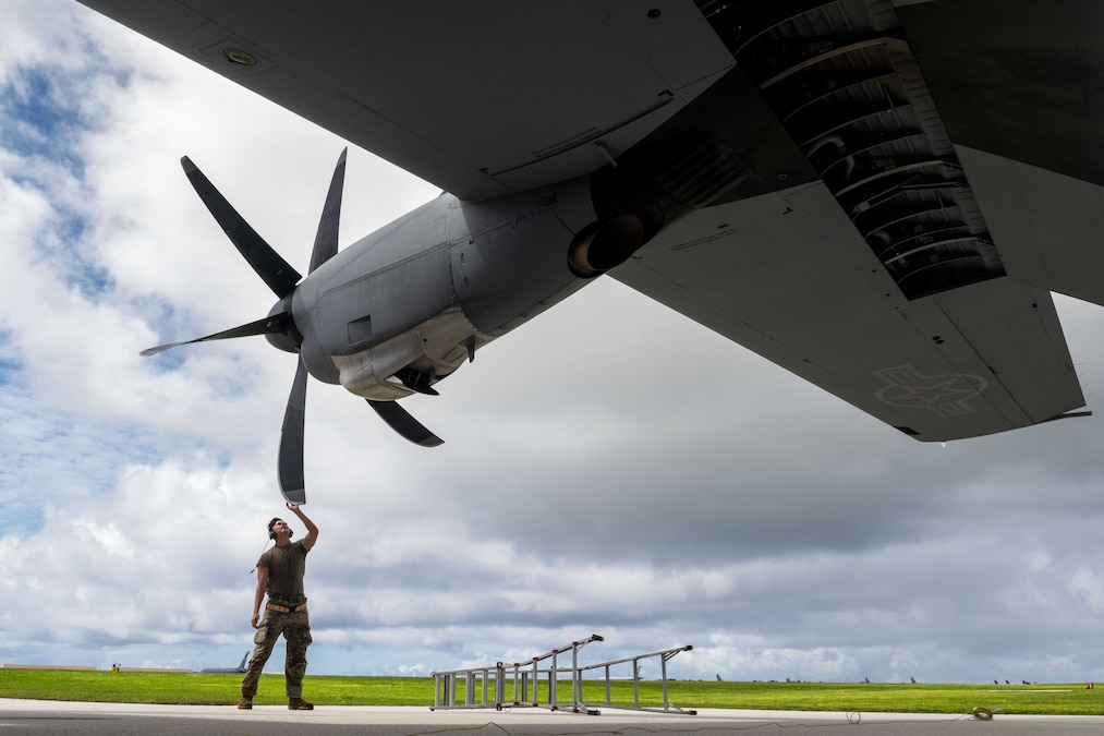 An airman reaches up to touch a propeller on an aircraft parked outside under cloudy skies.