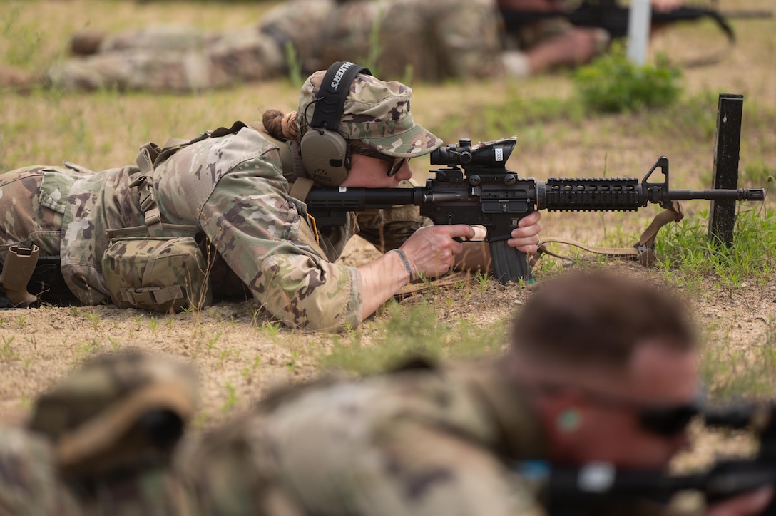 A soldier laying in the grass fires a service weapon while other soldiers are shown in the periphery doing the same.