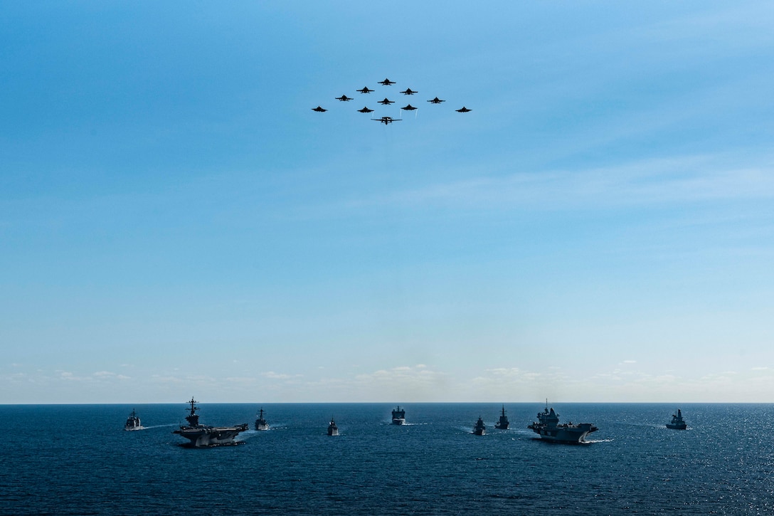 Eleven aircraft fly against blue skies in formation above nine ships sailing on the ocean.