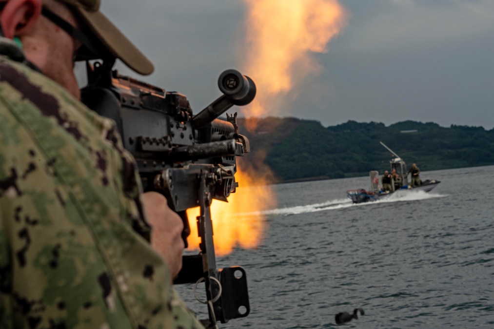 A sailor simulates firing a service weapon over the ocean as flames emerge and a small boat is shown in the distance.