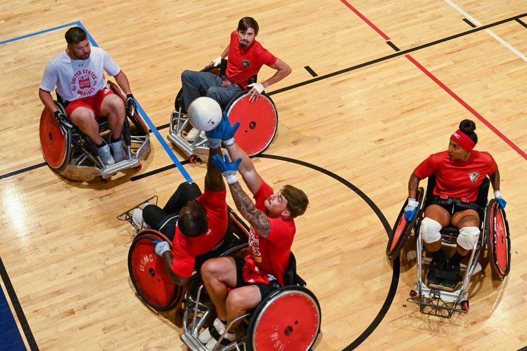 Five Marines in wheel chairs play a sport on a basketball court. Two Marines go up for a white ball.