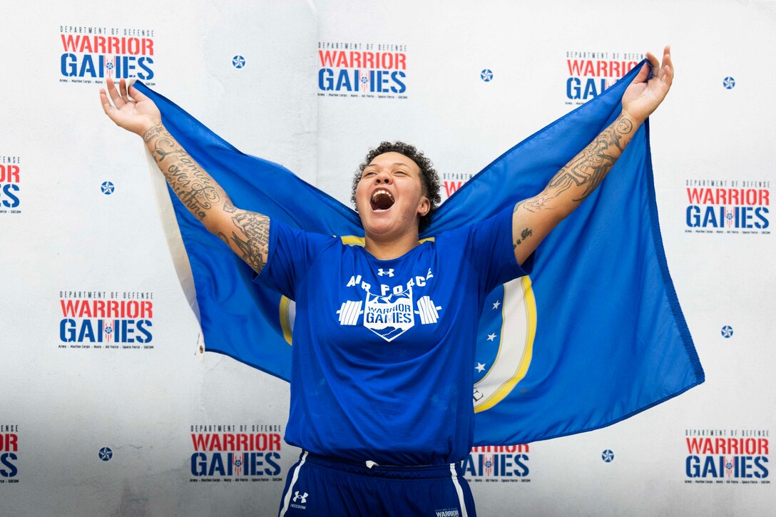 A woman wearing a blue shirt celebrates with a blue flag behind her back in front of a white background with the Warrior Games logo.