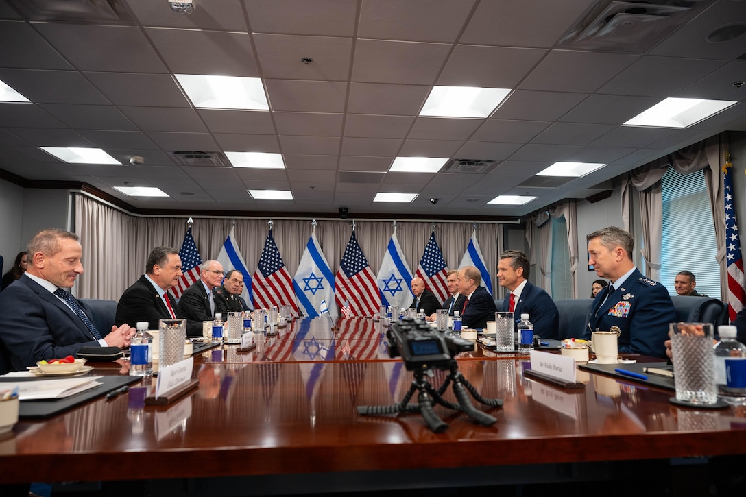 A group of people in suits sit around a wooden table with American and Israeli flags in the background.