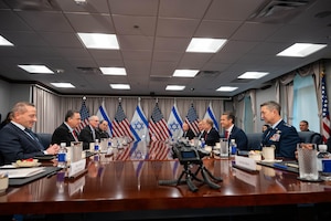 A group of people in suits sit around a wooden table with American and Israeli flags in the background.