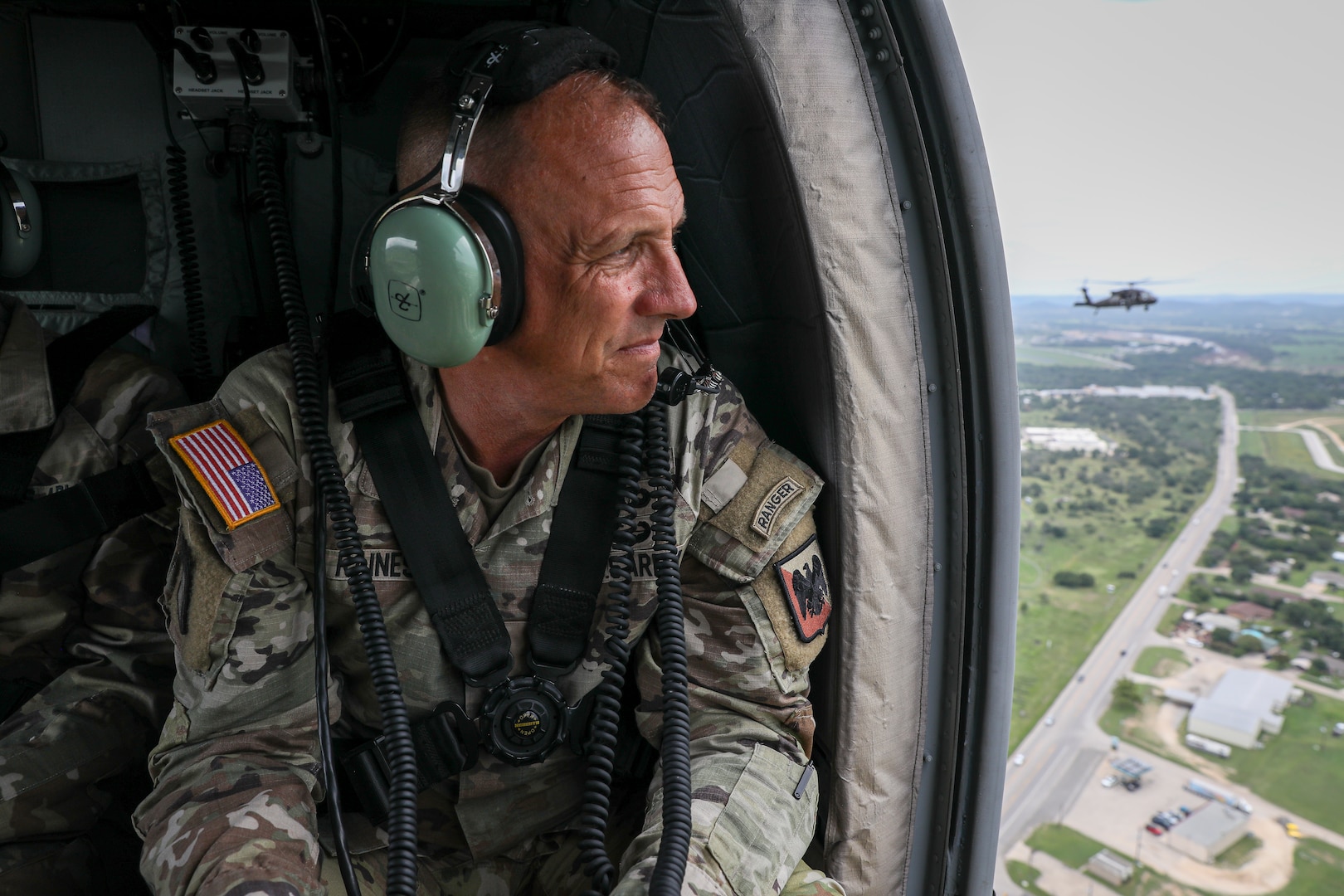 A man dressed in a military uniform looks out of the open door of a helicopter. There is another helicopter in the distance.
