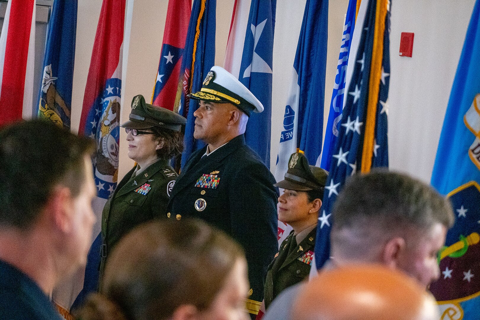 Photo of leaders participating in Transfer of Directorship ceremony from left to right, Col. Gina Adam, Rear Adm. Reginald Ewing, and Col. Elba Villacorta, standing at attention in front of flags with crowd in the foreground.