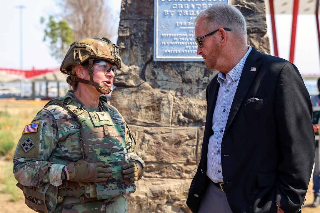 A soldier in gear speaks with a person in a blazer outside by a stone monument.