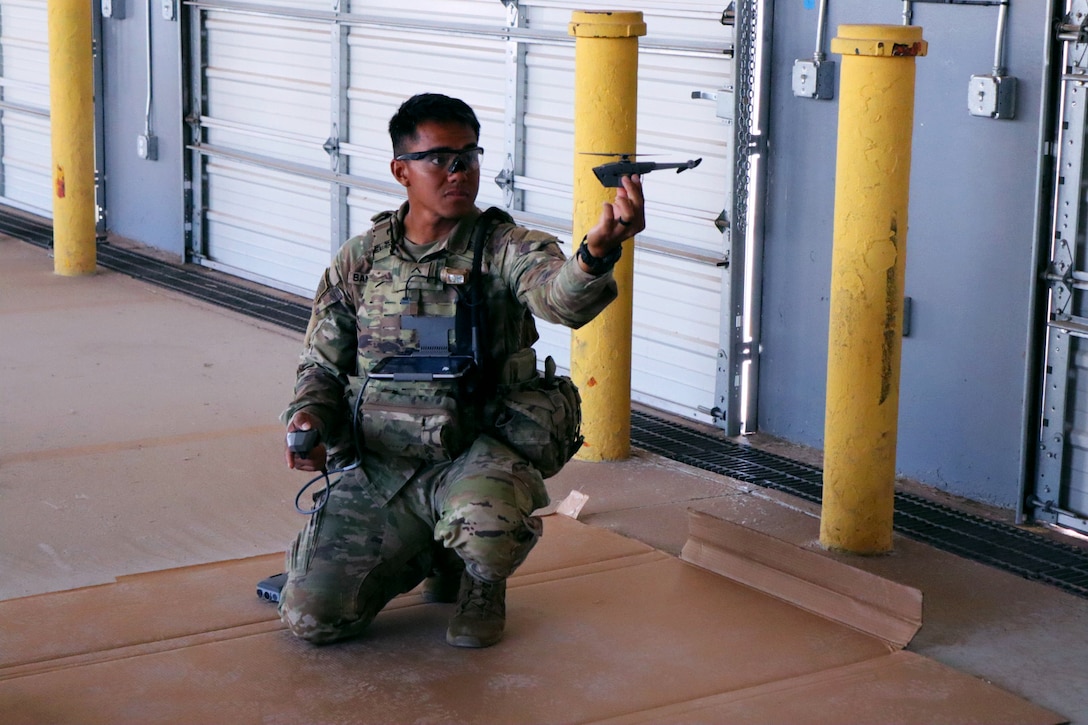A soldier in tactical gear holds out a small drone in one hand while crouching on the ground.