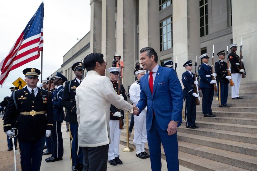 Two people wearing formal attire shake hands outside a building with steps and columns. Service members from each U.S. military branch stand in formation behind them; some hold ceremonial weapons, and one holds the American flag.