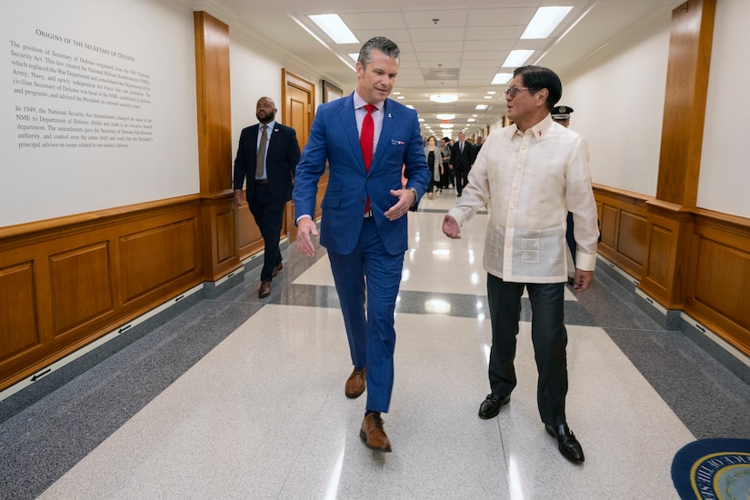 Two people wearing formal attire talk as they walk down a hallway with wood panels on the walls. A group of people is further behind them.