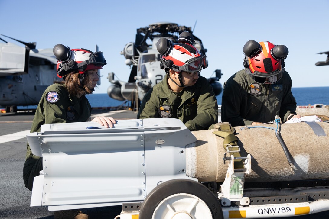 Three Marines work on a bomb while on the deck of a military ship during the day.