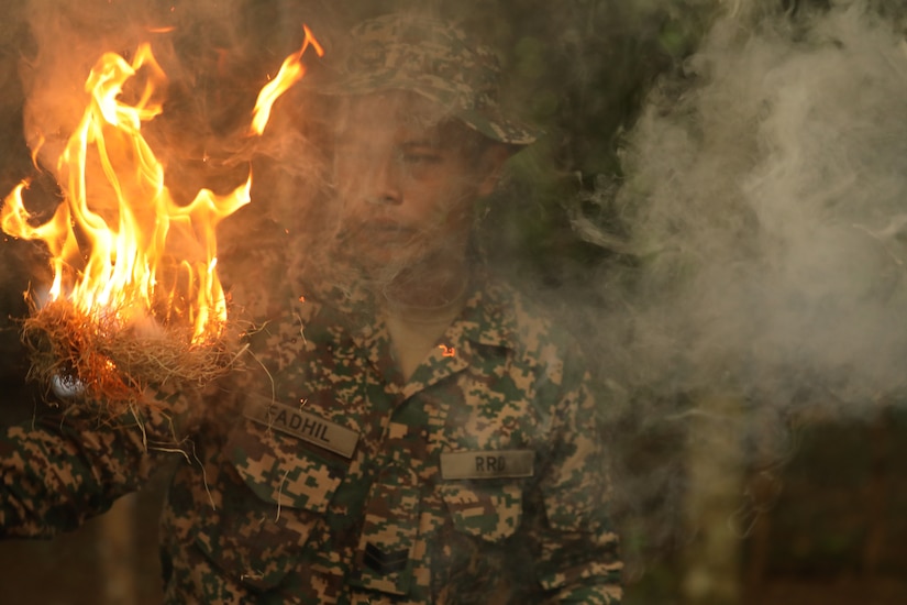 A Malaysian Army soldier demonstrates traditional fire-starting techniques, using natural materials such as dry grass and bark fibers along with a stone and knife to create the necessary tinder for a fire, an essential survival skill taught during jungle training to Australian and U.S. Army partners.