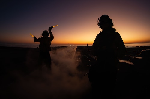 Two sailors stand on the deck of an aircraft carrier as one signals with lamps with minimal sunlight in the horizon.