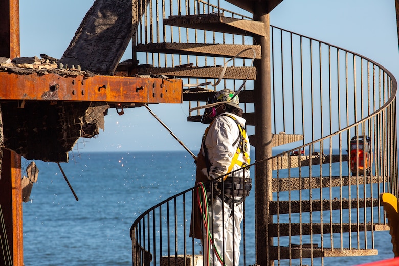 A man in safety clothing and gear stands on a spiral staircase of a burned-out building with the ocean in the background.