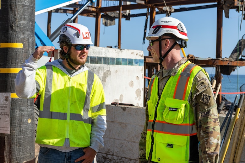 Two men in safety vests and hard hats talk with fire debris in the background