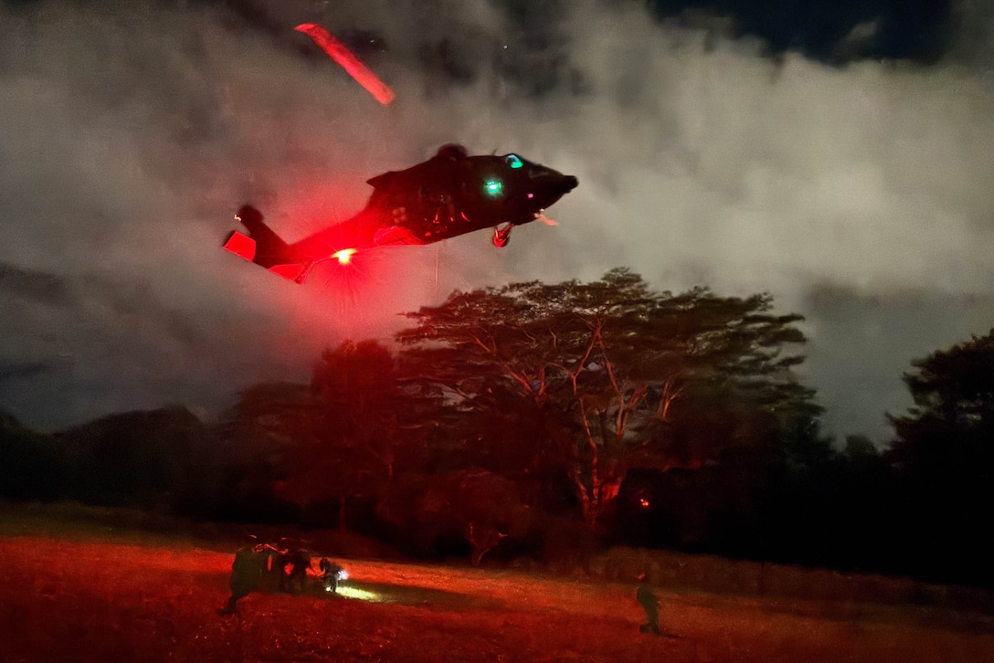 A helicopter hovers over a group of soldiers on a field with trees in the dark surrounded by red light.