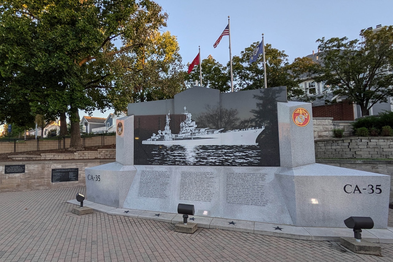 A large, stone monument depicting a ship and a list of names sits in a park-like setting.