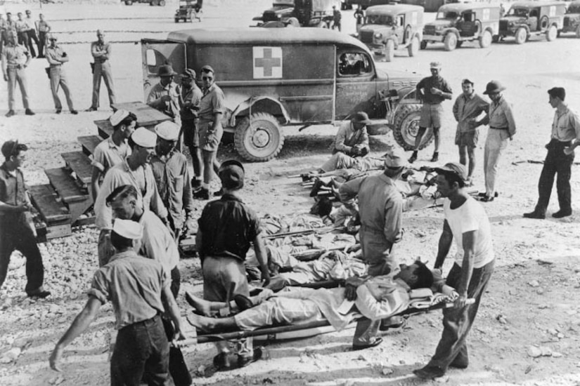 A group of people carries injured sailors on medical litters. There is an ambulance in the background.
