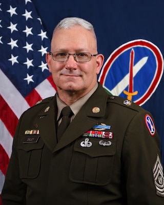 Army command sergeant major dressed in green service uniform is posting for a photo in front of the U.S. flag and an organizational flag that features a dark field and oval symbol in the middle with the Washington Monument and sword.