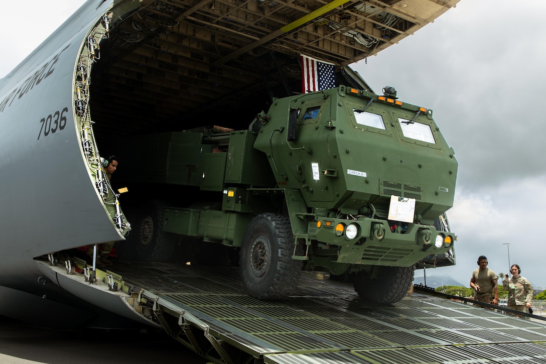 A green military vehicle rolls down the ramp of an aircraft while two service members watch from the side on the tarmac.