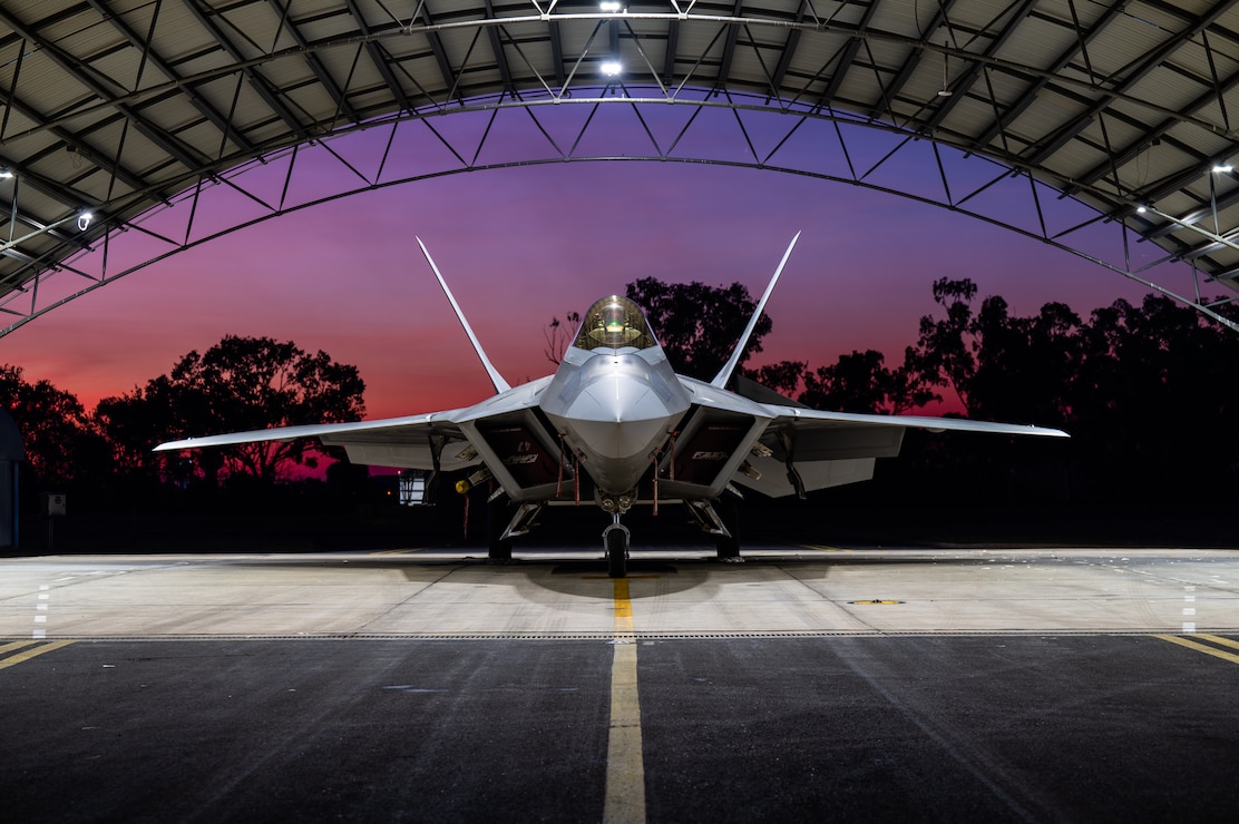 A U.S. Air Force F-22 Raptor assigned to the 199th Air Expeditionary Squadron sits beneath a hangar as the sun sets in Northern Territory, Australia, July 16, 2025, during Talisman Sabre 2025.