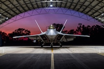 A U.S. Air Force F-22 Raptor assigned to the 199th Air Expeditionary Squadron sits beneath a hangar as the sun sets in Northern Territory, Australia, July 16, 2025, during Talisman Sabre 2025.
