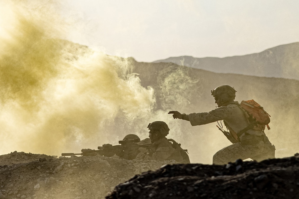 Marines and sailors take cover behind a pile of rocks while yellow smoke billows.