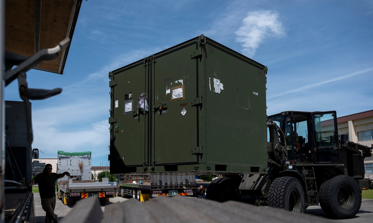 A military member moves a large green container on a forklift into a truck.