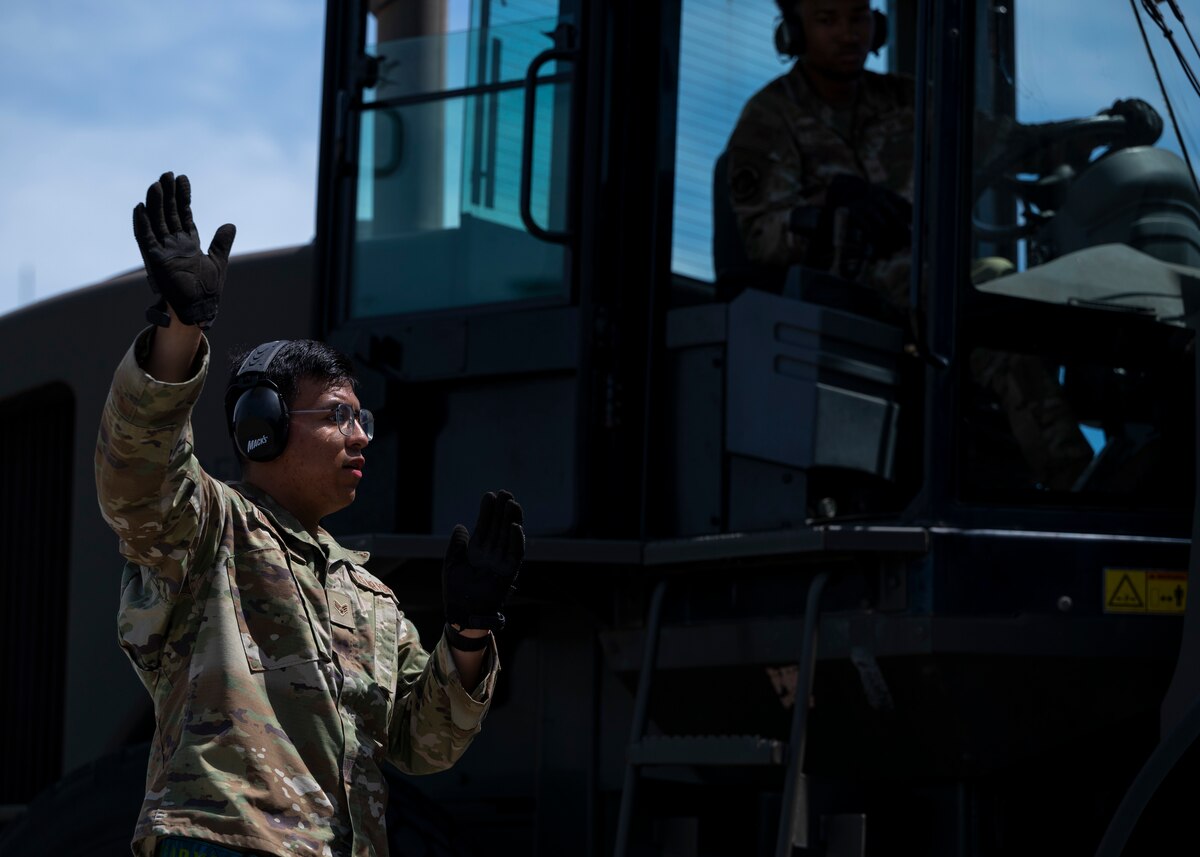 A military member signals a member driving a large forklift.