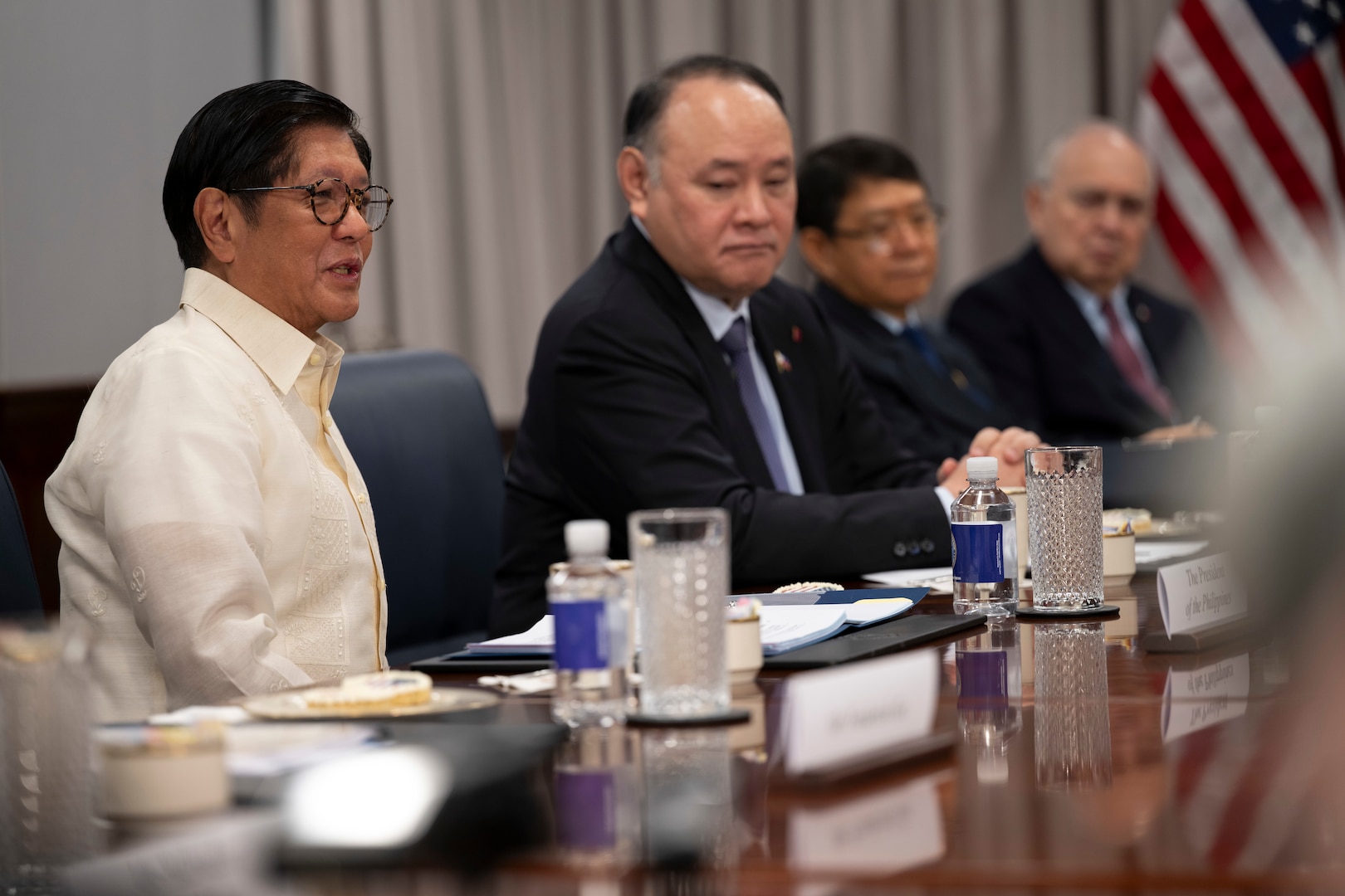 A person wearing formal attire sits at a wooden desk and speaks while three other people in similar attire sit to his left.