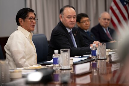 A person wearing formal attire sits at a wooden desk and speaks while three other people in similar attire sit to his left.