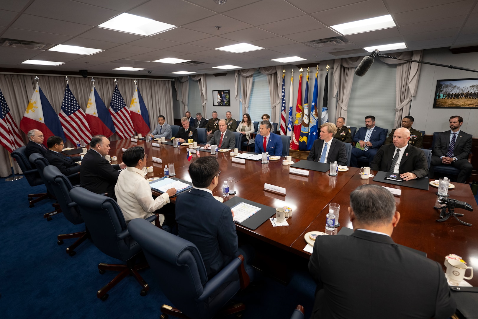 A group of people dressed in formal attire sit around a large wooden desk and talk while others sit in chairs behind them along a wall in a conference room. To the left are alternating American and Philippine flags.