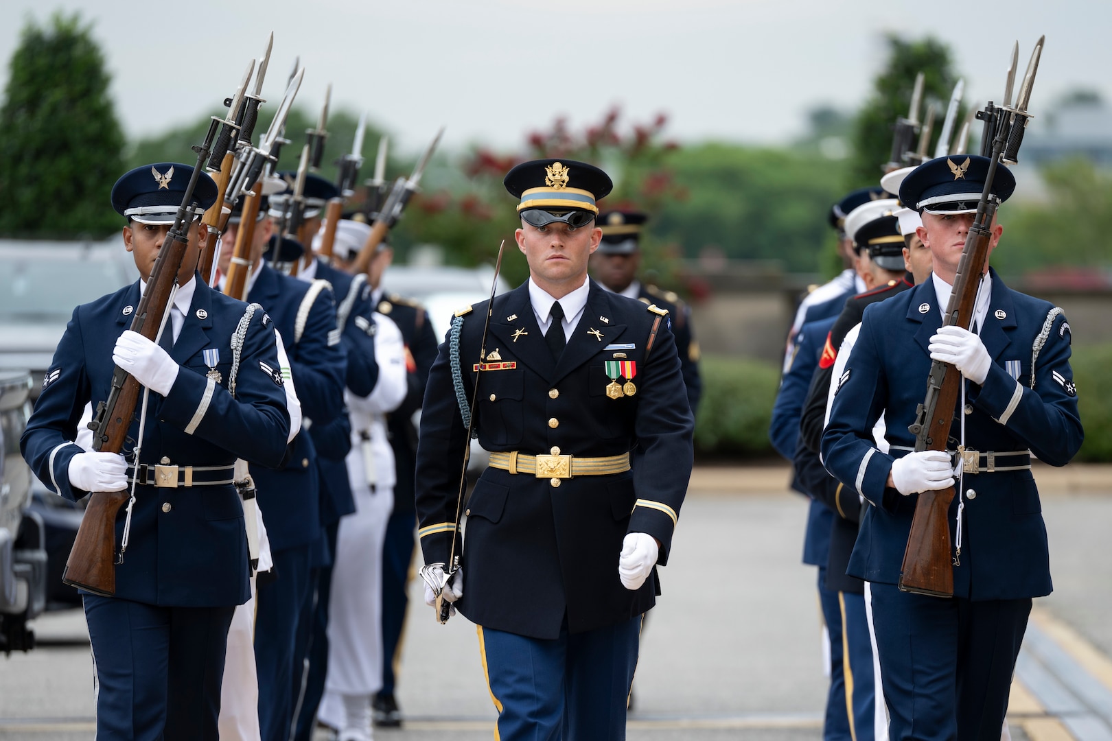 A group of U.S. service members wearing formal military uniforms march in formation while holding ceremonial weapons.