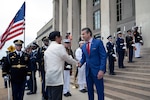 Two people wearing formal attire shake hands outside a building with steps and columns. Service members from each U.S. military branch stand in formation behind them; some hold ceremonial weapons, and one holds the American flag.