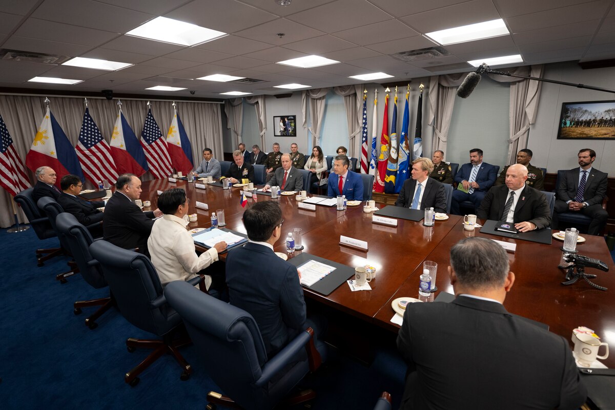 A group of people dressed in formal attire sit around a large wooden desk and talk while others sit in chairs behind them along a wall in a conference room. To the left are alternating American and Philippine flags.