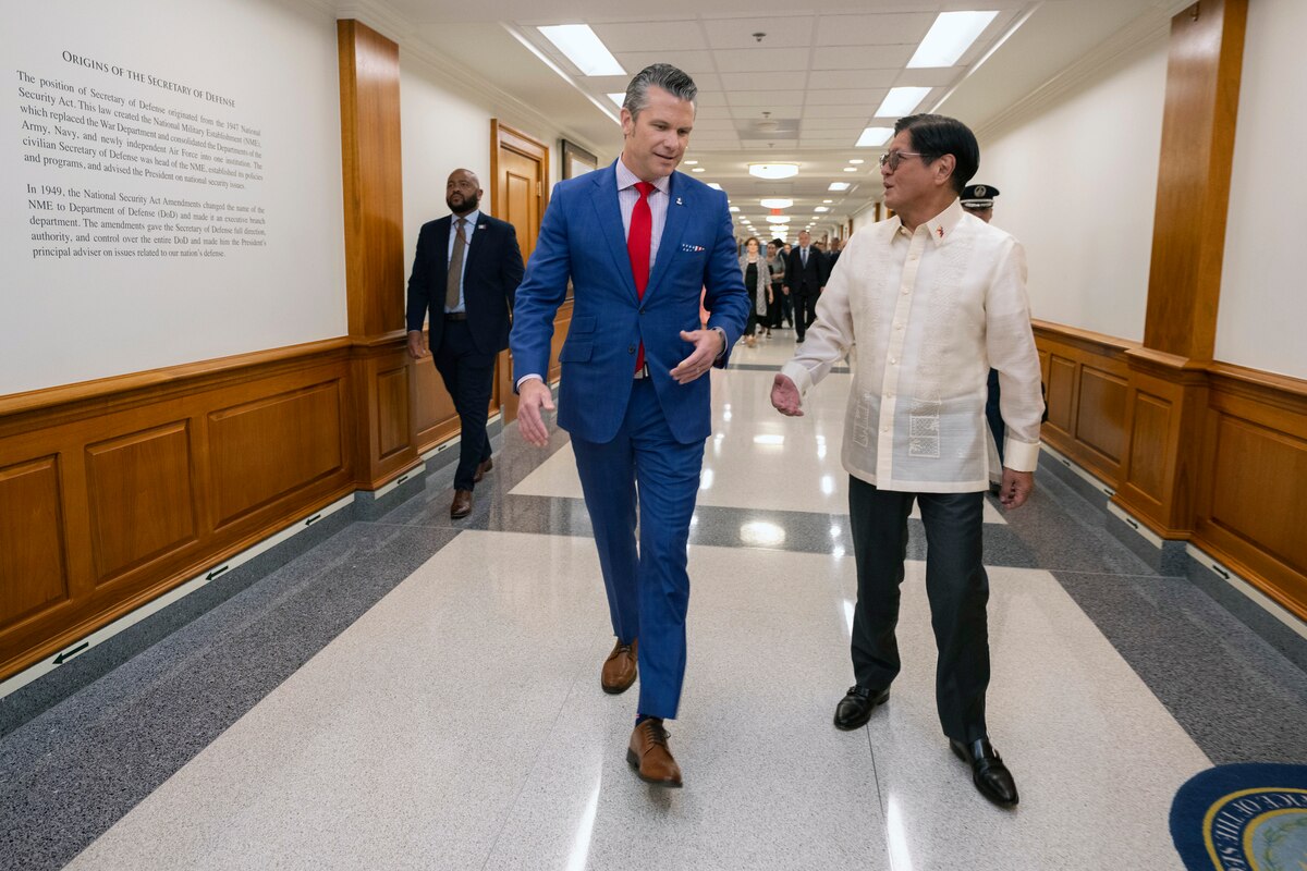 Two people wearing formal attire talk as they walk down a hallway with wood panels on the walls. A group of people is further behind them.