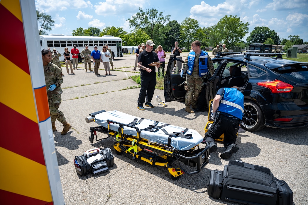 A large group of people watch as first responders set up a stretcher and check a passenger in a black car