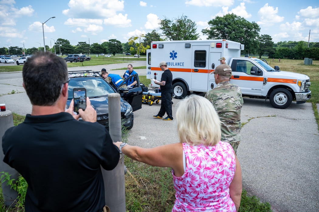 Multiple people can be seen between an ambulance and wrecked car while some people stand in the foreground with backs to viewer